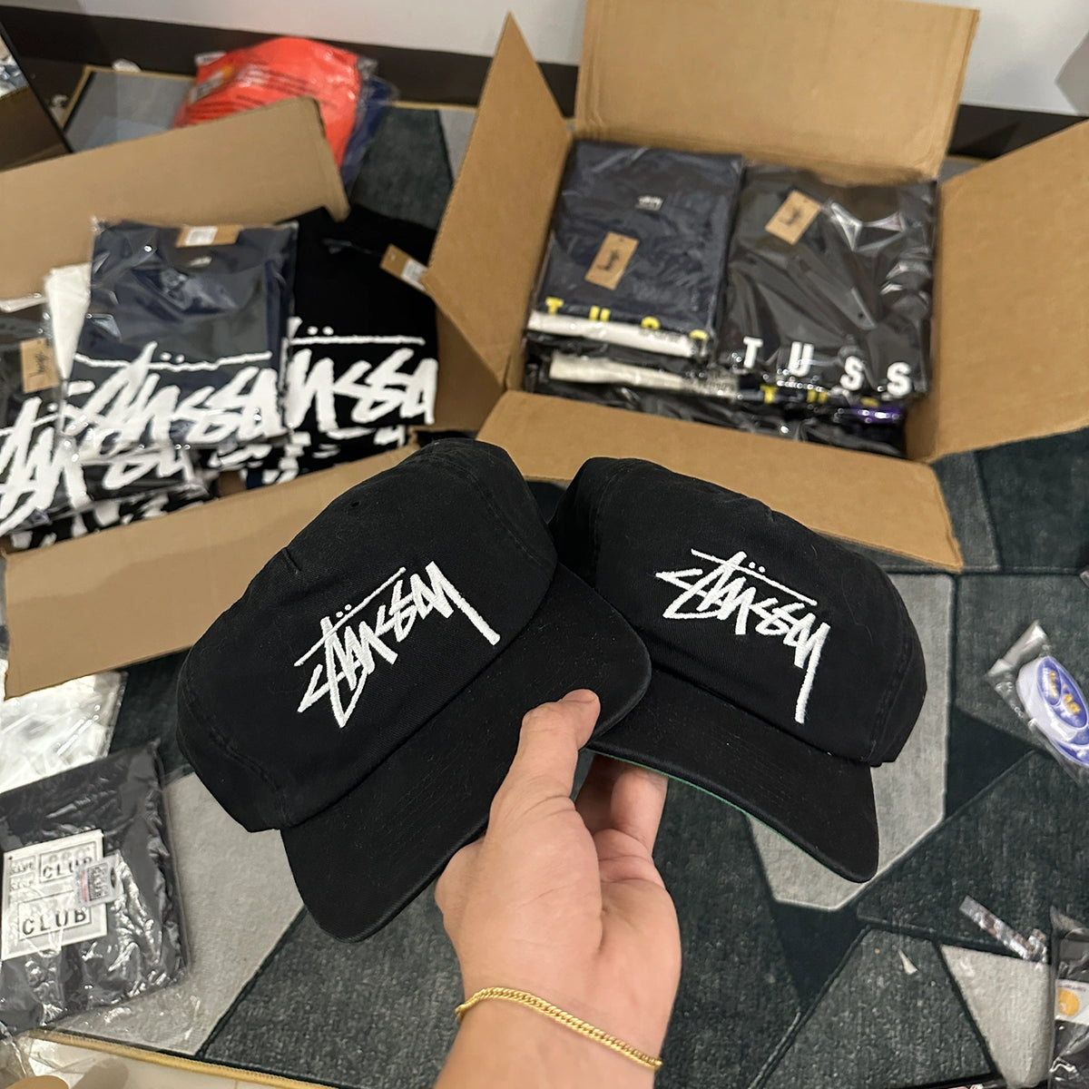 Two black Stussy snapback hats held by a hand, with open cardboard boxes of Stussy clothing in the background.