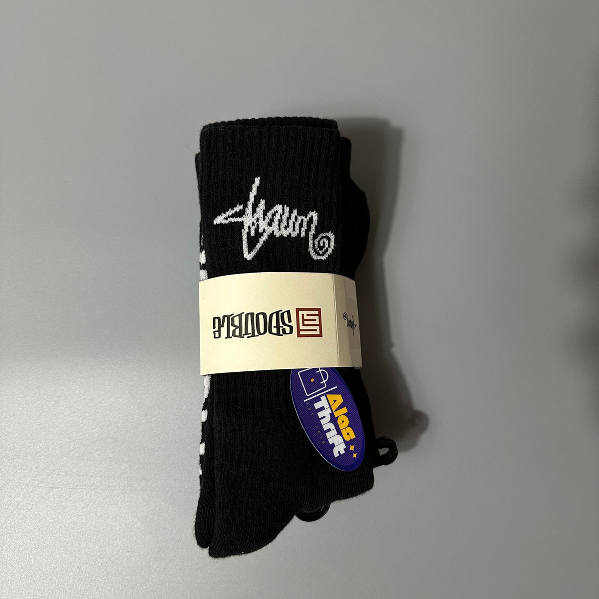 A pair of black knit socks bundled with a paper band, featuring a white script logo and a blue 'Alad Theft' tag, set against a plain gray background.